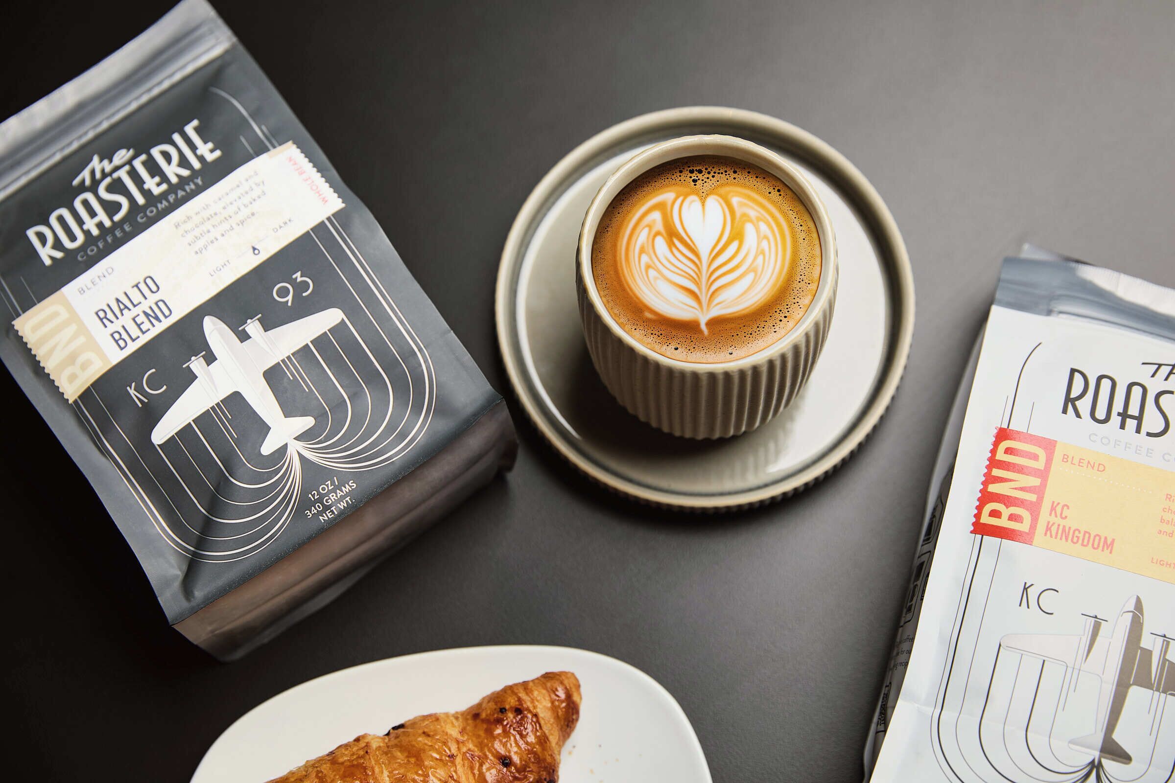 A coffee cup with latte art, a coffee bag, and a croissant on a plate.