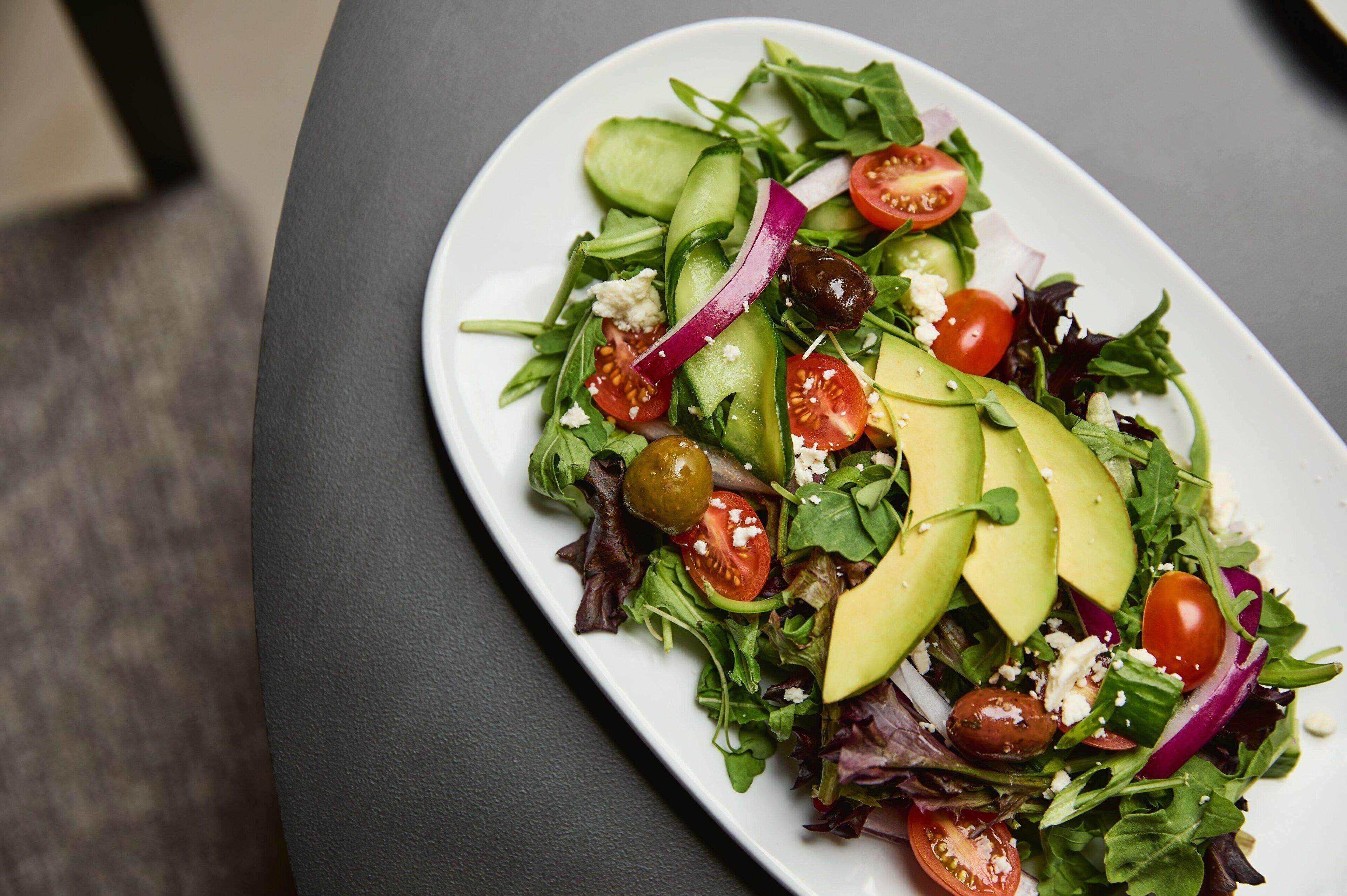 Uma salada de folhas verdes variadas com pepino, tomate, cebola roxa e fatias de abacate.