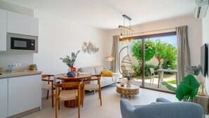 Una sala moderna con un sofá, una mesa de comedor y sillas. Hay una zona de cocina con microondas y armarios. Un amplio ventanal ofrece vistas a zonas verdes.