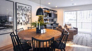 Un espace repas moderne avec une table ronde en bois, des chaises noires et un luminaire suspendu. Un grand tableau encadré orne le mur, et une bibliothèque contient divers objets décoratifs.