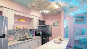 Cozinha moderna com armários brancos, geladeira de aço inoxidável, micro-ondas e fogão. Há uma ilha branca com banquetas e um bolo decorativo.