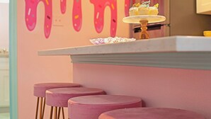 Uma bancada de cozinha com um bolo em um suporte, parede rosa com um design de tinta escorrendo, e três banquetas rosa.