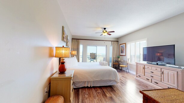 A hotel room with a bed, a television, a desk with a lamp, a window with blinds, and a balcony with a chair. - Summit 0409a (Miramar Beach)