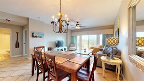 A spacious living area with a dining table, chairs, a sofa, and a TV. There is a ceiling fan, a chandelier, and a large window providing natural light. - Summit 0409 (Miramar Beach)
