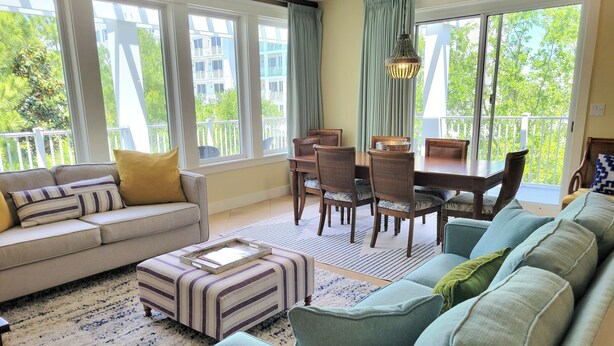 Ein geräumiges Wohnzimmer mit einer Couch, Hocker und einem Essbereich mit Stühlen und Tisch. Große Fenster bieten einen Blick ins Freie.