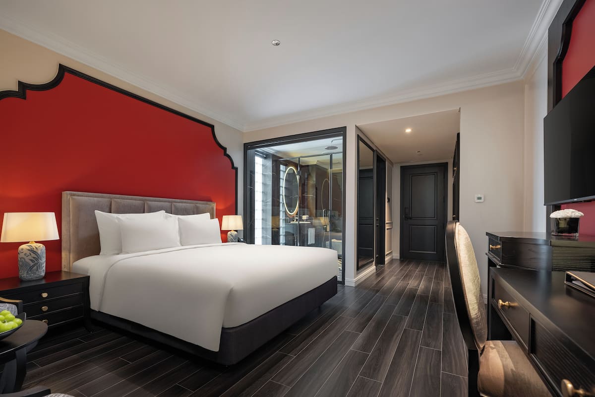 Habitación de hotel moderna con una cama grande, piso de madera oscura, una pared de acento roja y un escritorio con televisión.