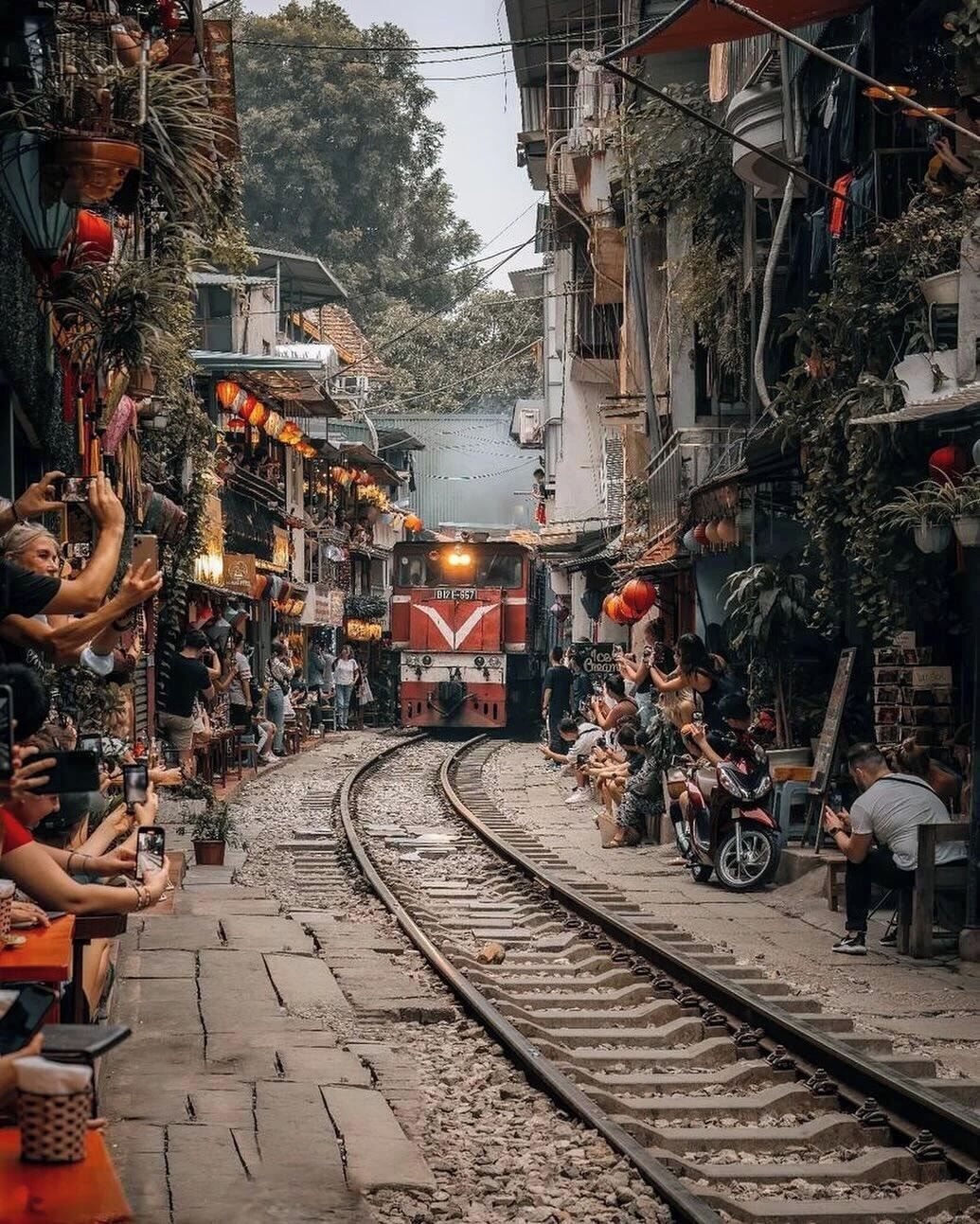 A narrow street with a train track running through it, flanked by buildings with balconies and outdoor seating areas.