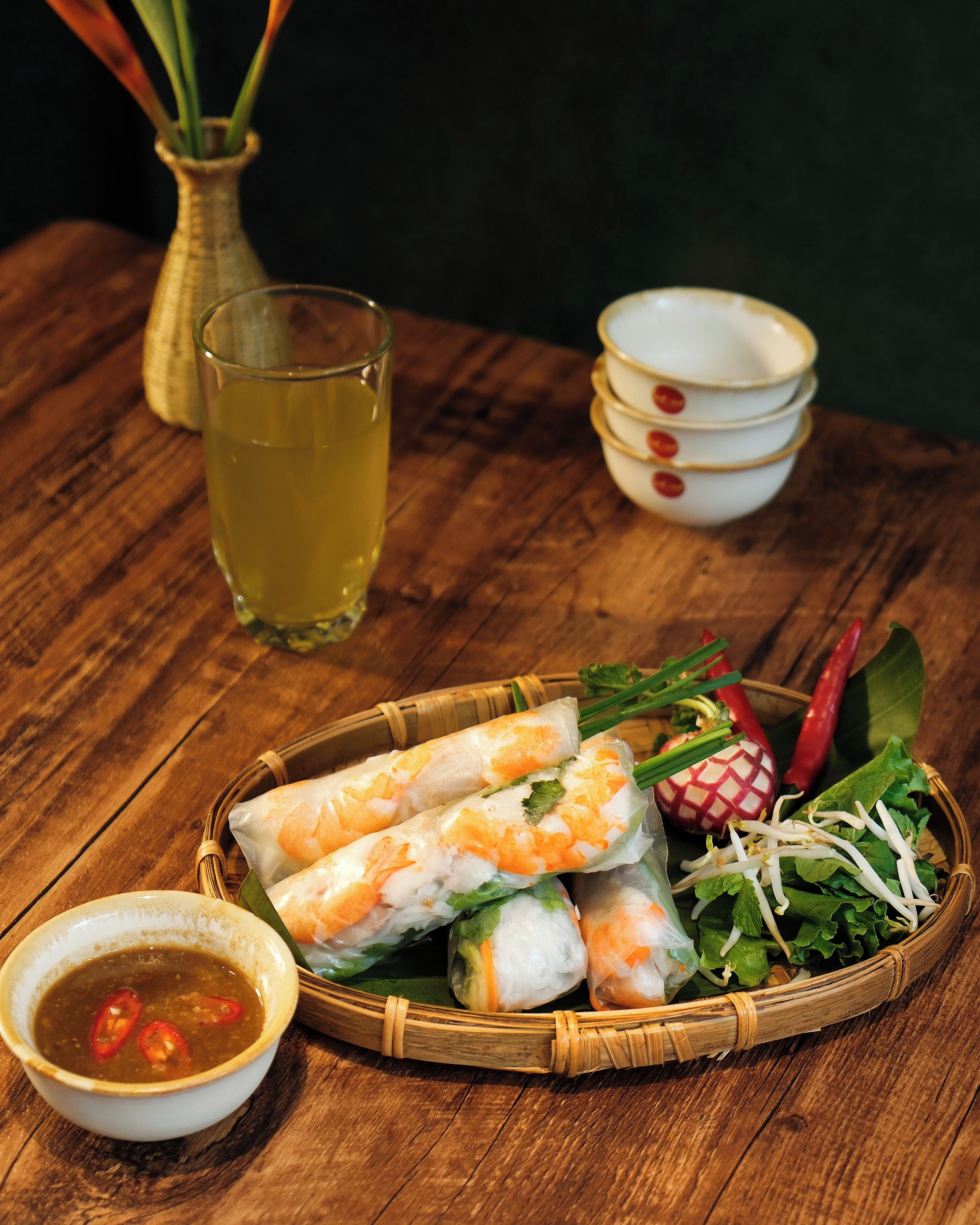Ein Holztisch mit einem Korb vietnamesischer Sommerrollen, einem Glas gelber Flüssigkeit und einer Schale Dip-Soße.