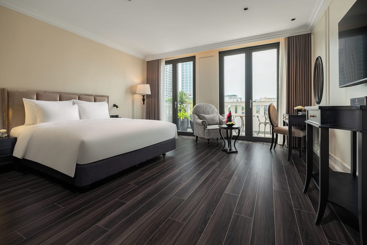 Habitación de hotel con una cama grande, un escritorio con silla, una mesita de noche con lámpara y vistas a la ciudad a través de amplios ventanales.