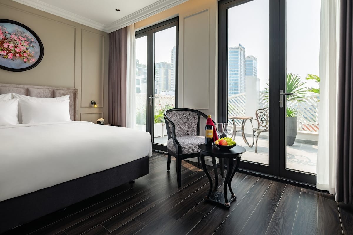 Habitación de hotel con una cama grande, una mesa de comedor con una silla y un balcón con vistas a la ciudad.