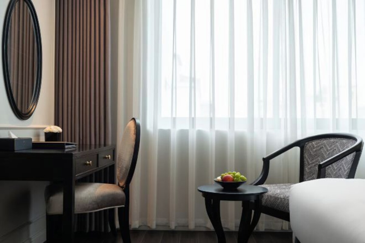A hotel room with a bed, two chairs, a small table with fruit, a desk with a lamp, and a large mirror.