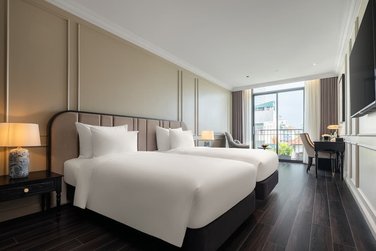 Habitación de hotel con dos camas, un escritorio, una silla y un ventanal con vistas a la ciudad.