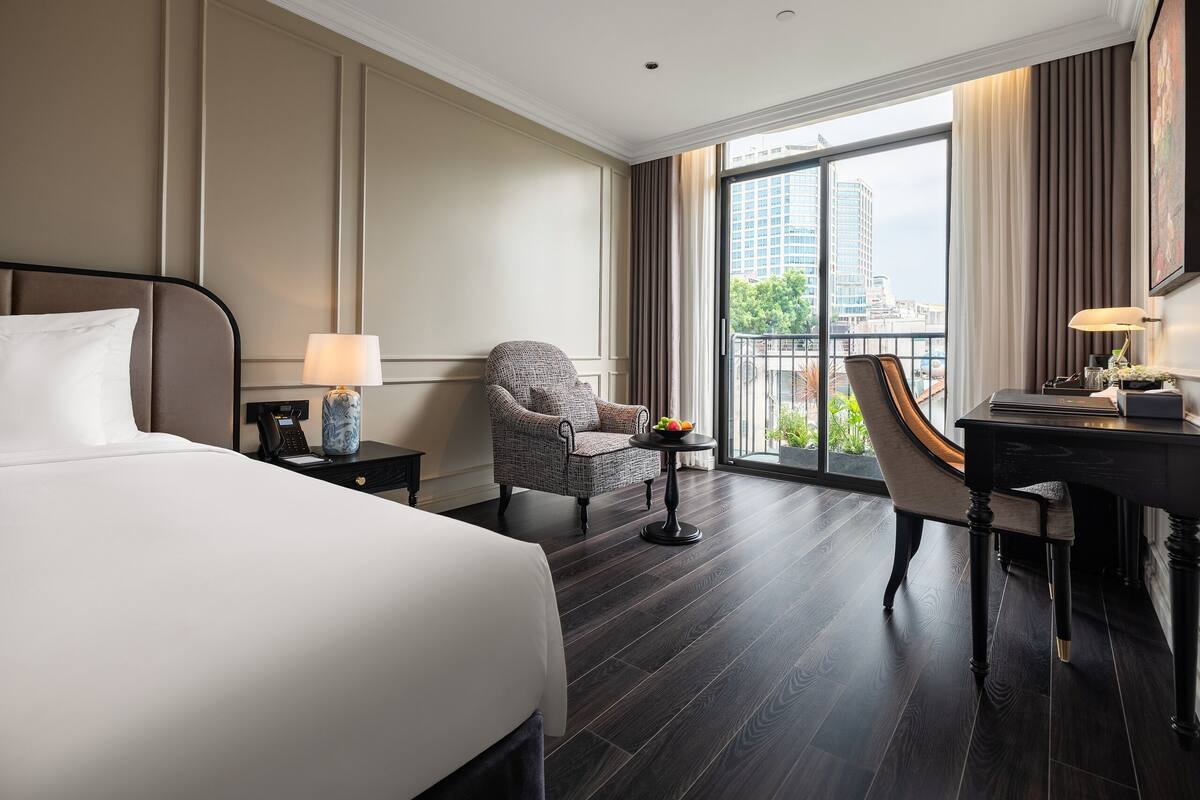 Habitación de hotel con una cama grande, un escritorio, una silla y vistas de la ciudad a través de un amplio ventanal.