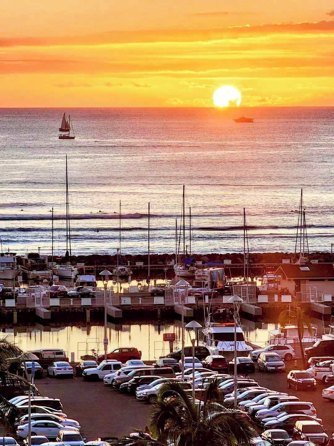 Een jachthaven met afgemeerde boten, een ondergaande zon op de achtergrond en een parkeerplaats met auto's.