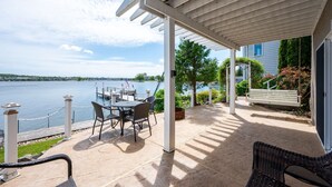 Une terrasse couverte, dotée d’une table et de chaises, qui offre une vue sur un lac avec un quai et un drapeau.