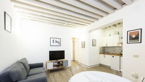 Een moderne woonkamer met een grijze bank, een televisie op een houten dressoir, een marmeren tafel, en een keuken op de achtergrond.