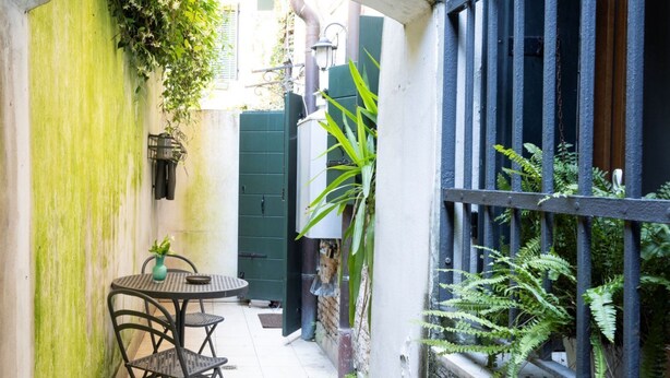 Een smal terras buiten, met een ronde tafel en stoelen, een potplant en een groene poort.