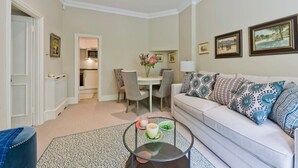 A living room with a sofa, a coffee table, a dining area with chairs, and framed pictures on the wall. - Spacious Studio Flat, Holland Park (Greater London)