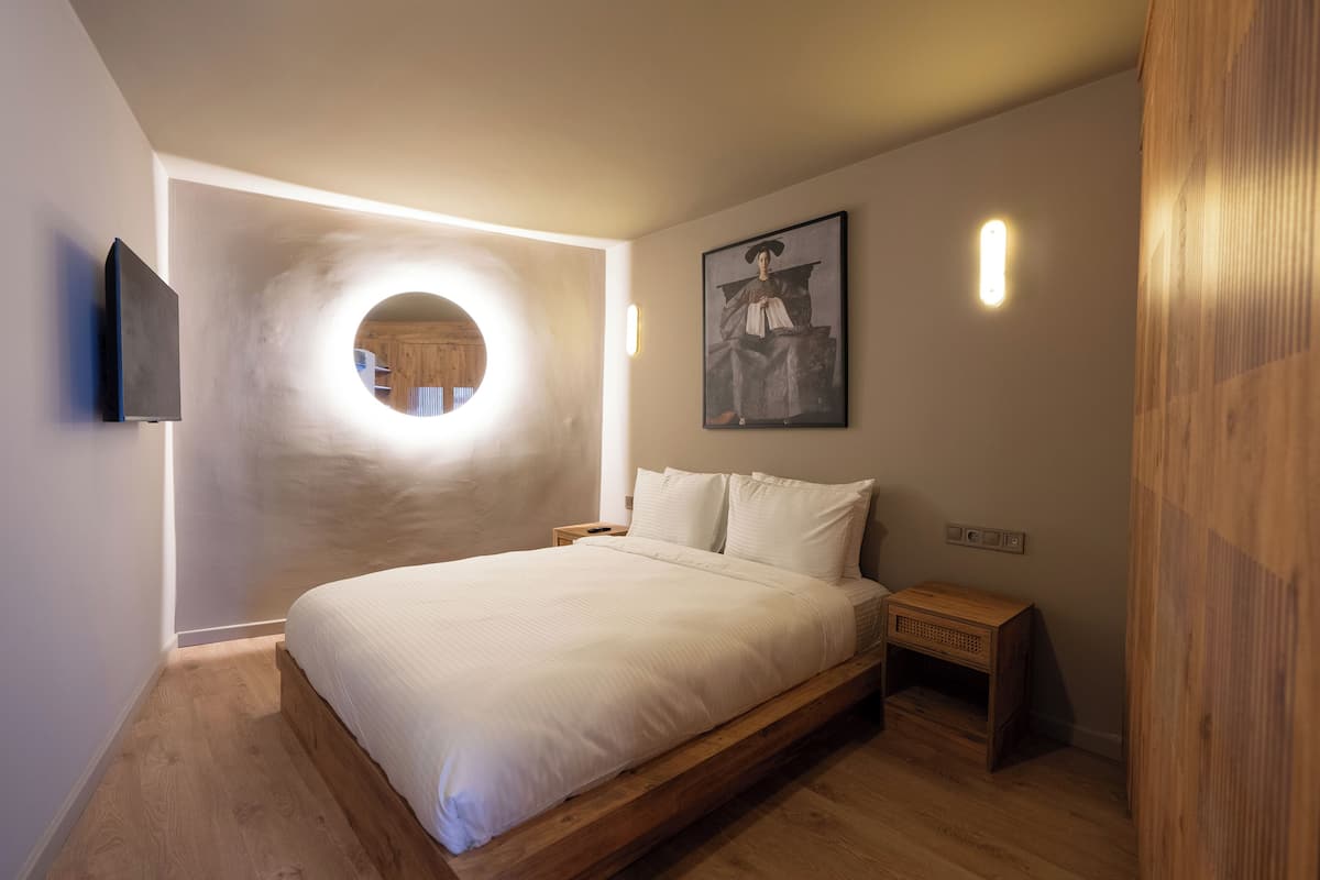 A bedroom with a circular skylight, a bed with white bedding, a nightstand, a wooden floor, and a painting on the wall.