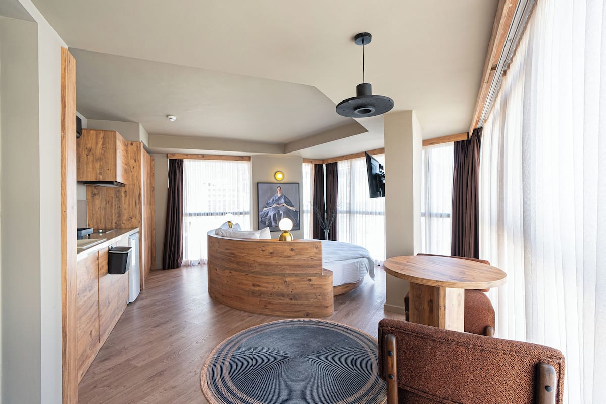 A modern hotel room with a curved wooden desk, a round dining table, a comfortable sofa, and a large bed.