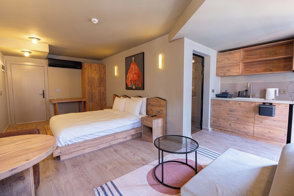 A modern bedroom with a wooden bed, bedside tables, a small round table, and a kitchenette area with wooden cabinets and a sink.