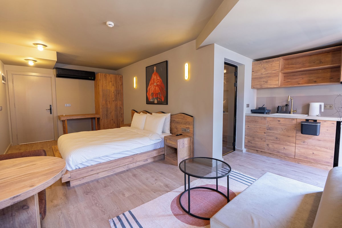 A modern bedroom with a wooden bed, bedside tables, a small round table, and a kitchenette area with wooden cabinets and a sink.