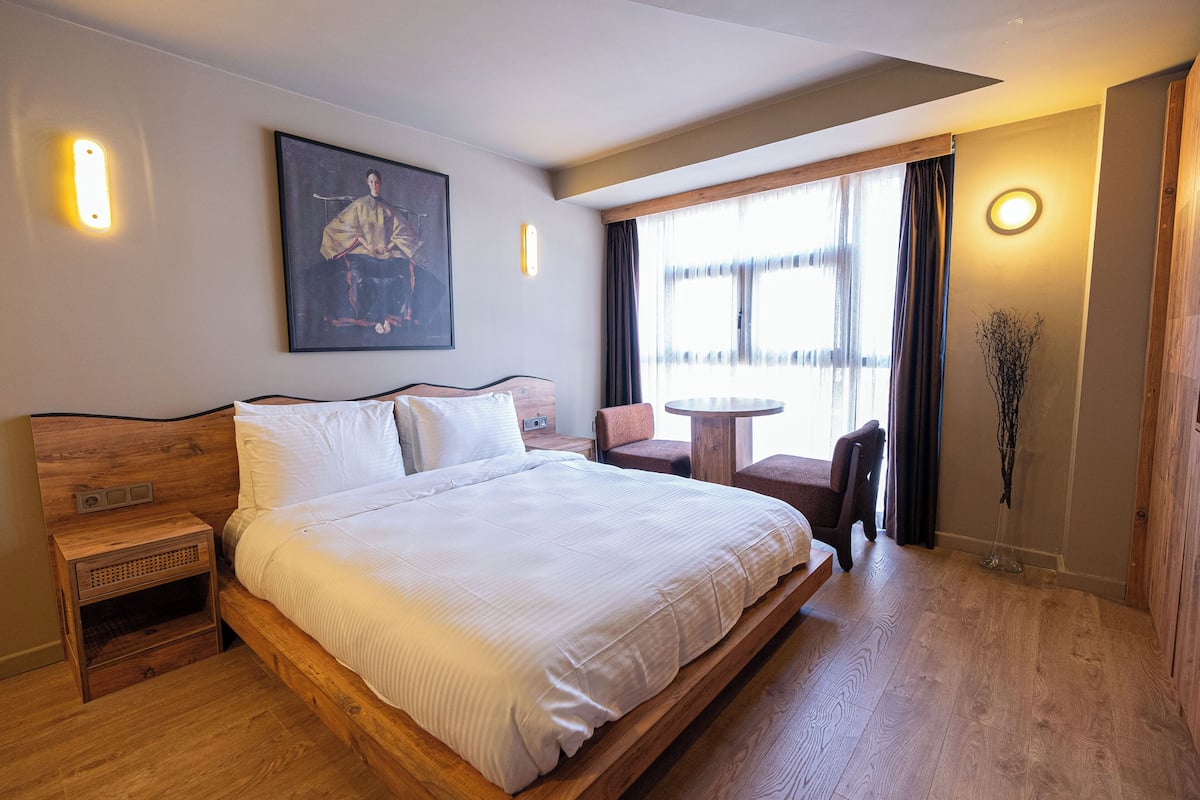 A spacious bedroom with a large bed, a wooden nightstand, a small table, and a chair. There is a window with curtains, a painting on the wall, and a vase with branches.