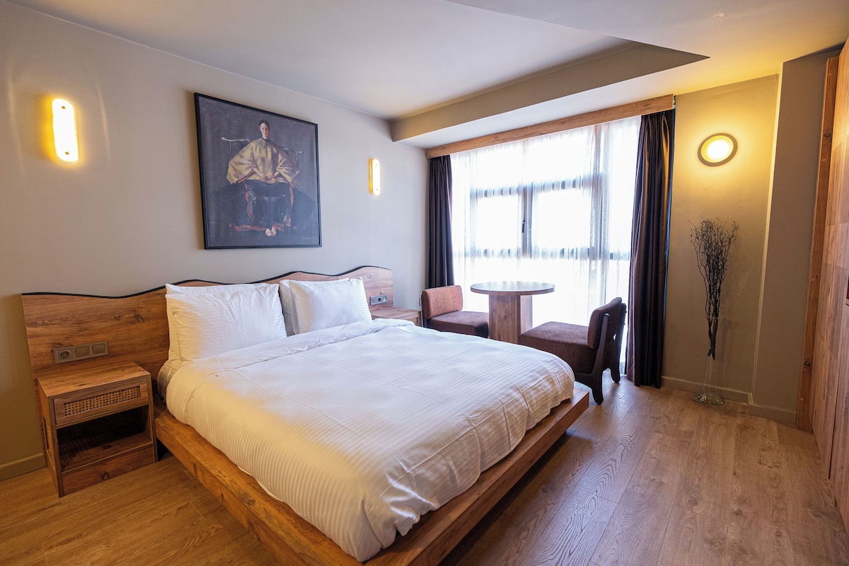 A spacious bedroom with a large bed, a wooden nightstand, a small table, and a chair. There is a window with curtains, a painting on the wall, and a vase with branches.