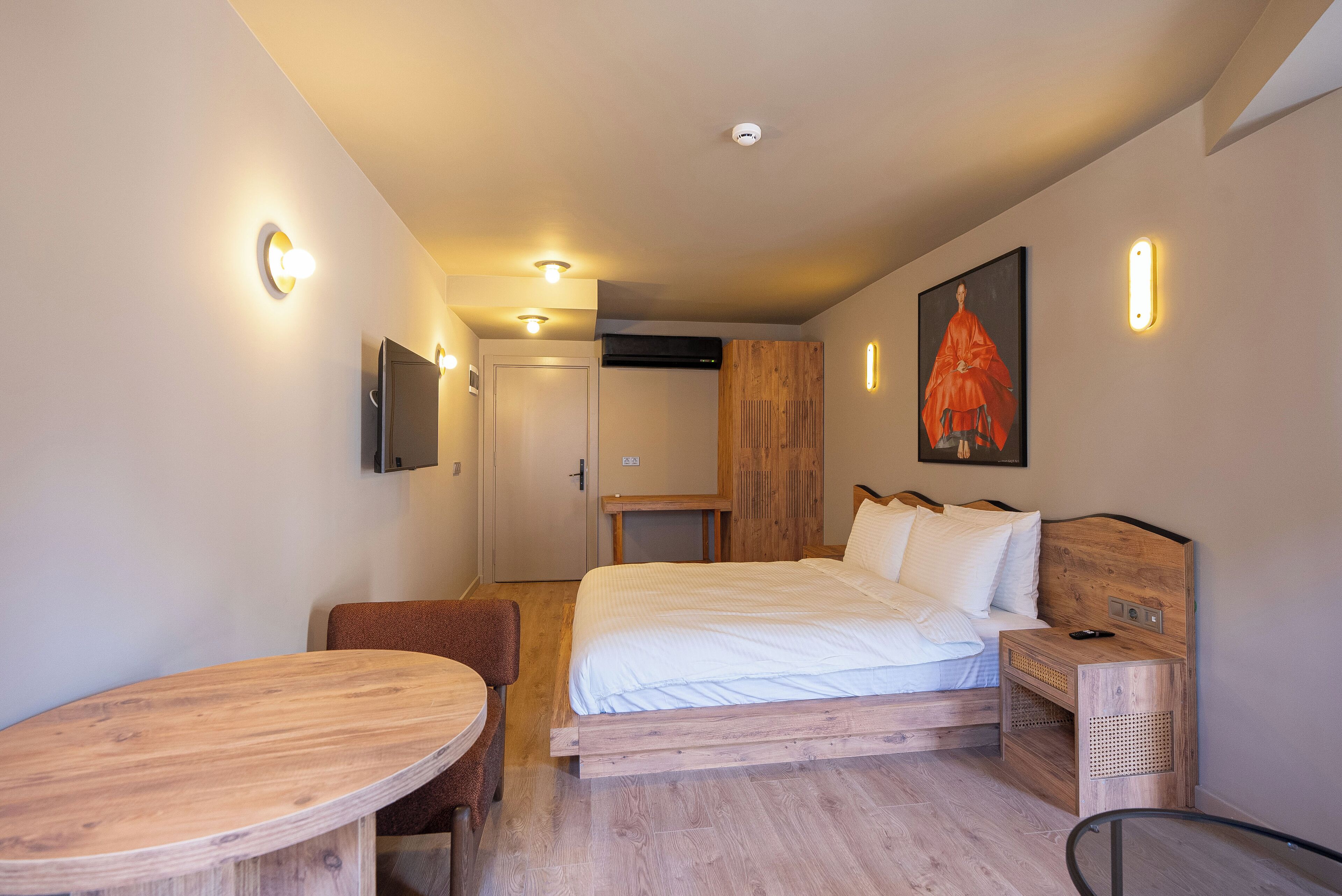 A modern hotel room with a wooden bed, a small wooden table, a TV mounted on the wall, and a painting.