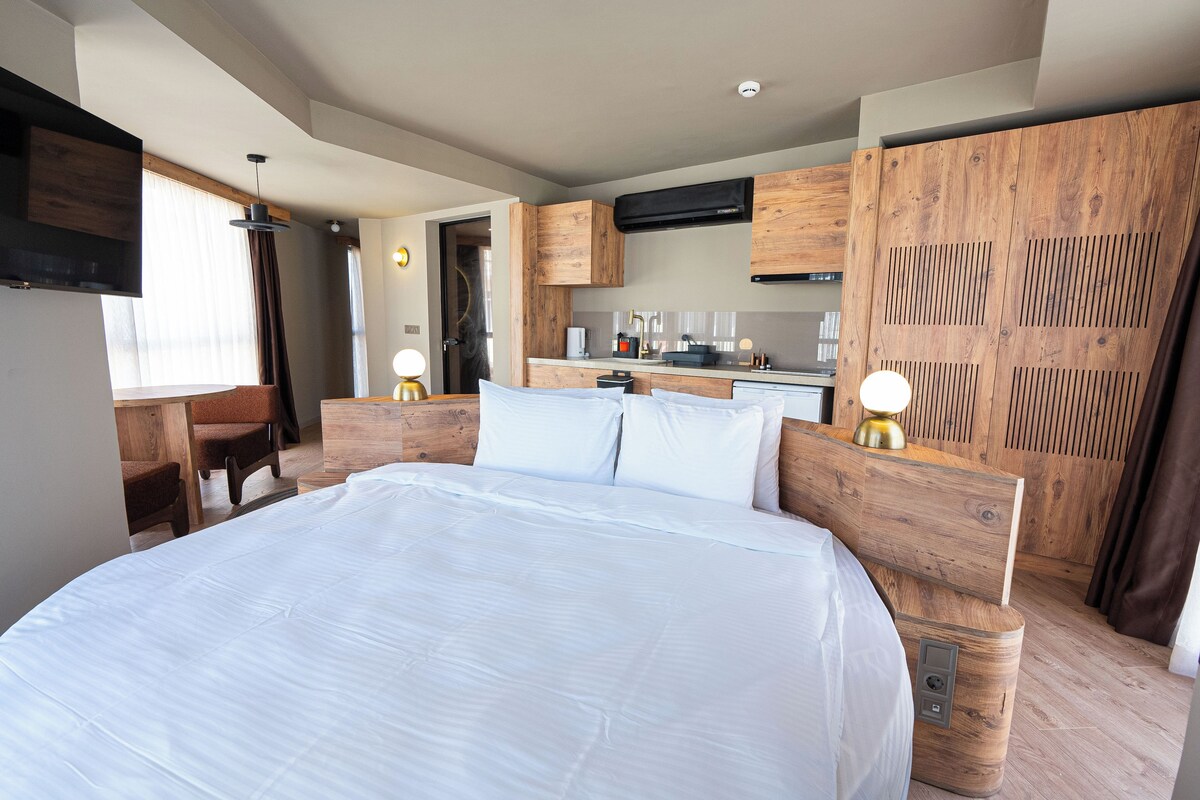 A modern hotel room with a large bed, wooden headboard, a desk, and a kitchenette.