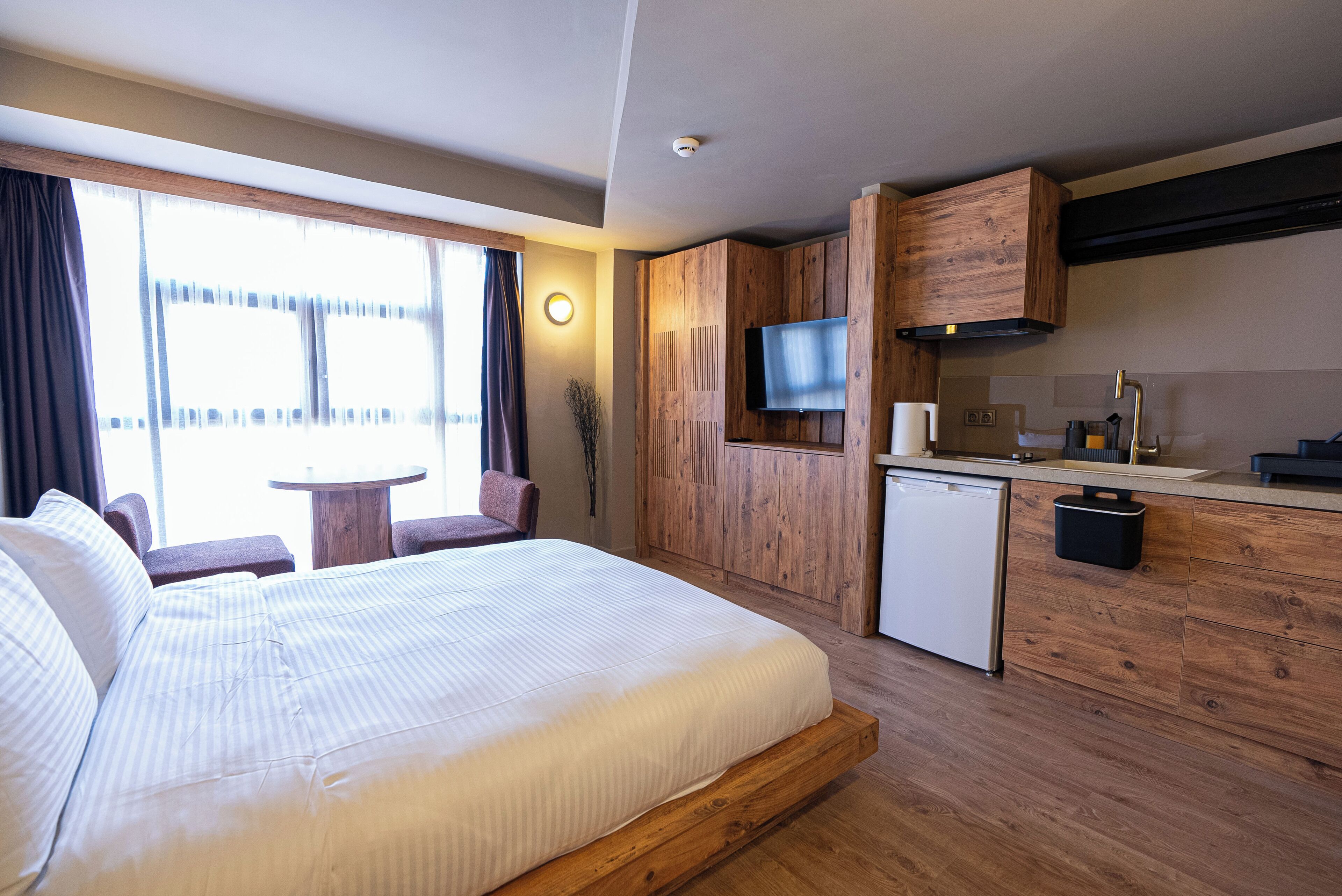Quarto de hotel com cama, pequeno espaço de cozinha, televisão e janela com cortinas.