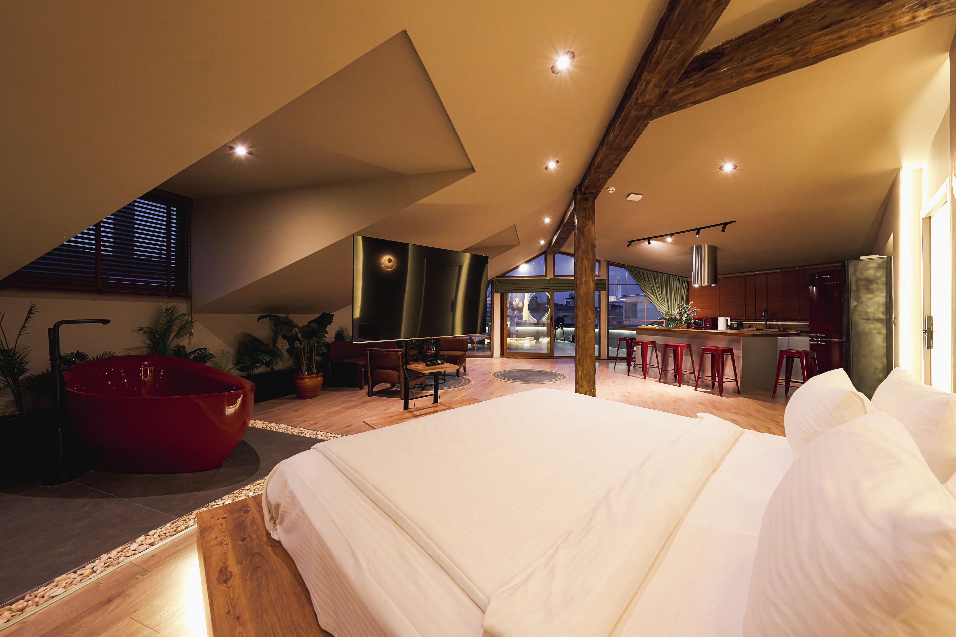 A modern, spacious bedroom with a large bed, a red bathtub, and a kitchen area with stools.