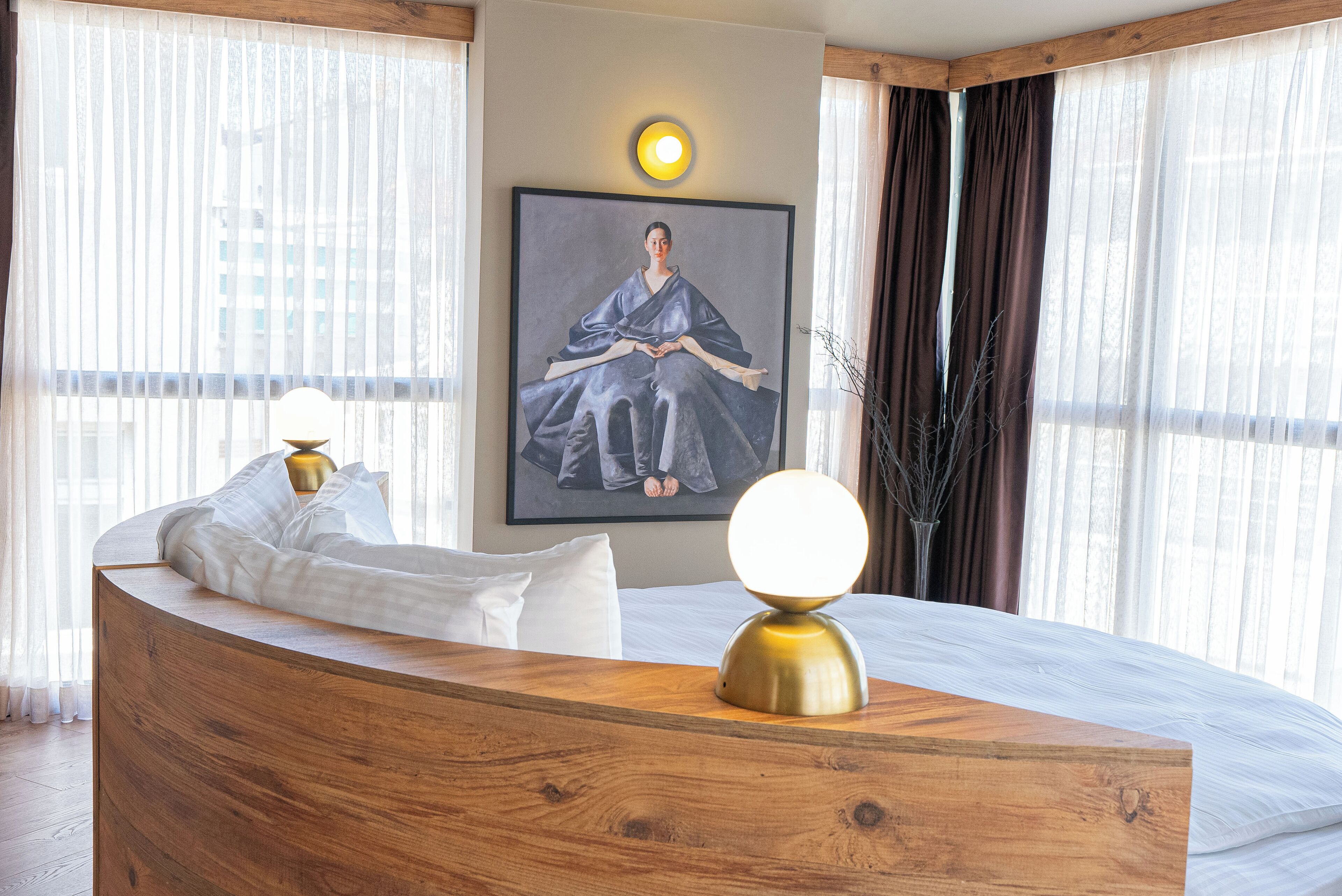 A modern hotel room with a wooden reception desk, a bed with white linens, a framed artwork on the wall, and a large window with sheer curtains.