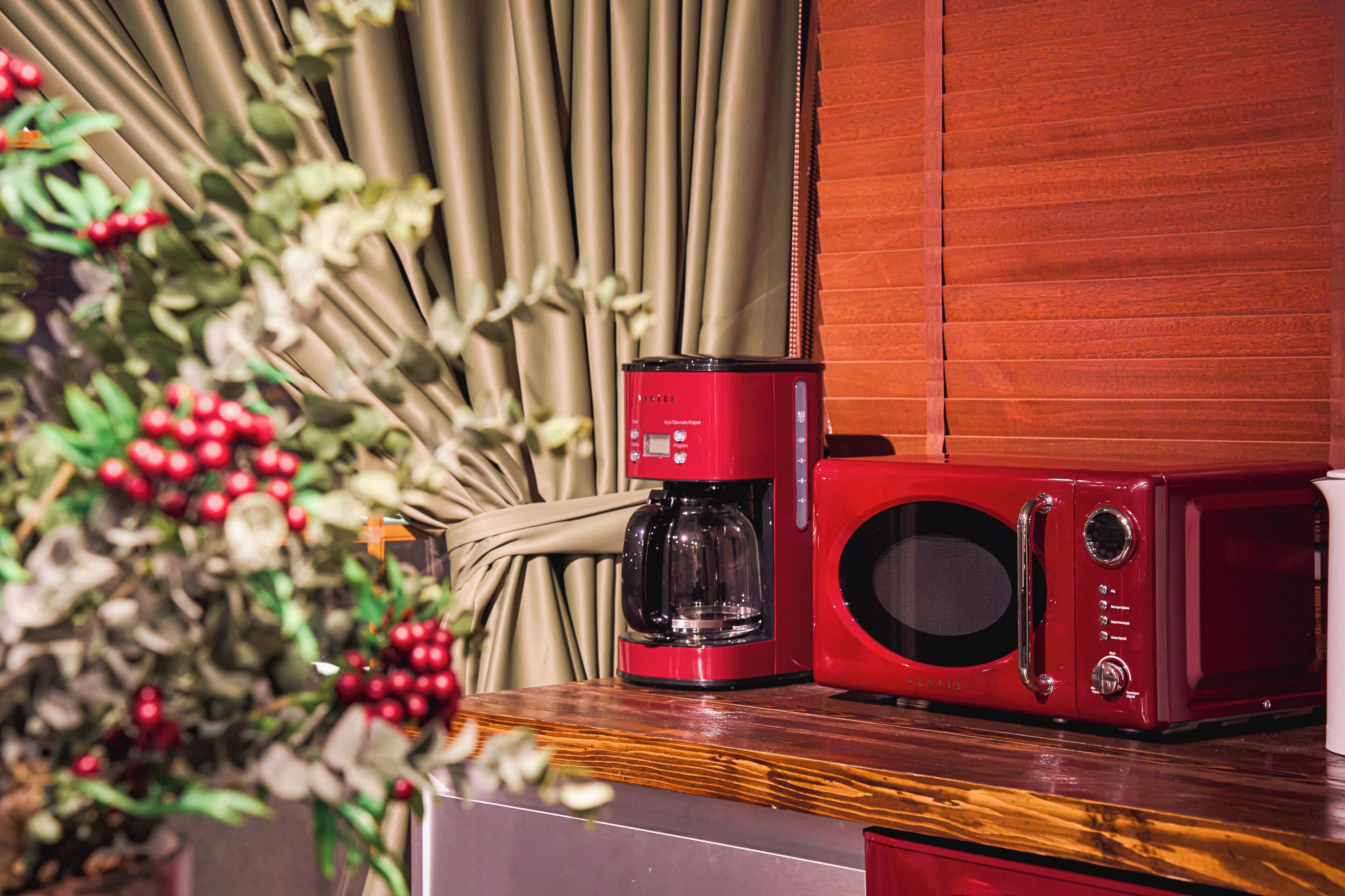 A red coffee maker and microwave on a wooden surface with a curtain and plant in the background.