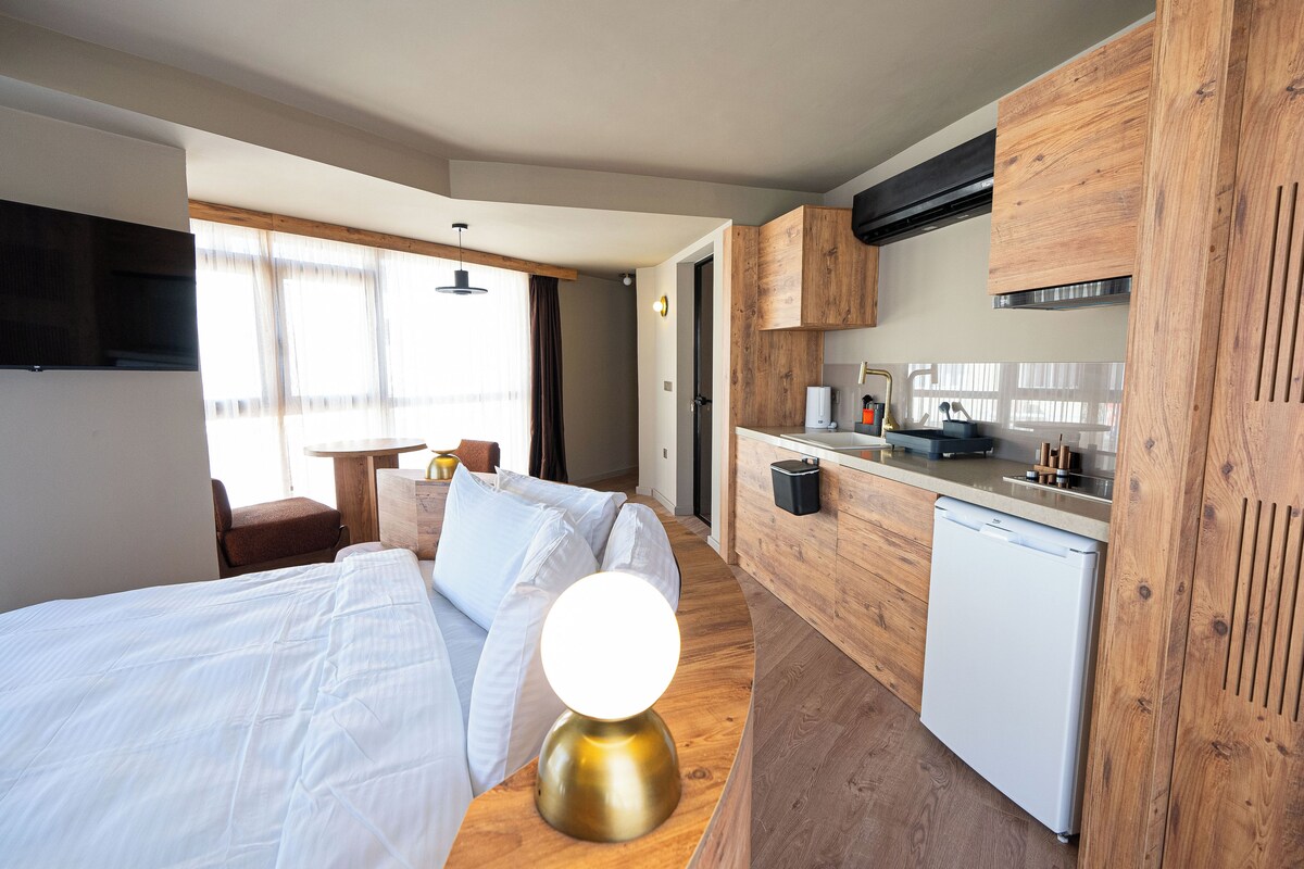 A compact hotel room with a bed, bedside lamp, wooden cabinets, a small kitchenette, and a view of the outside through large windows.