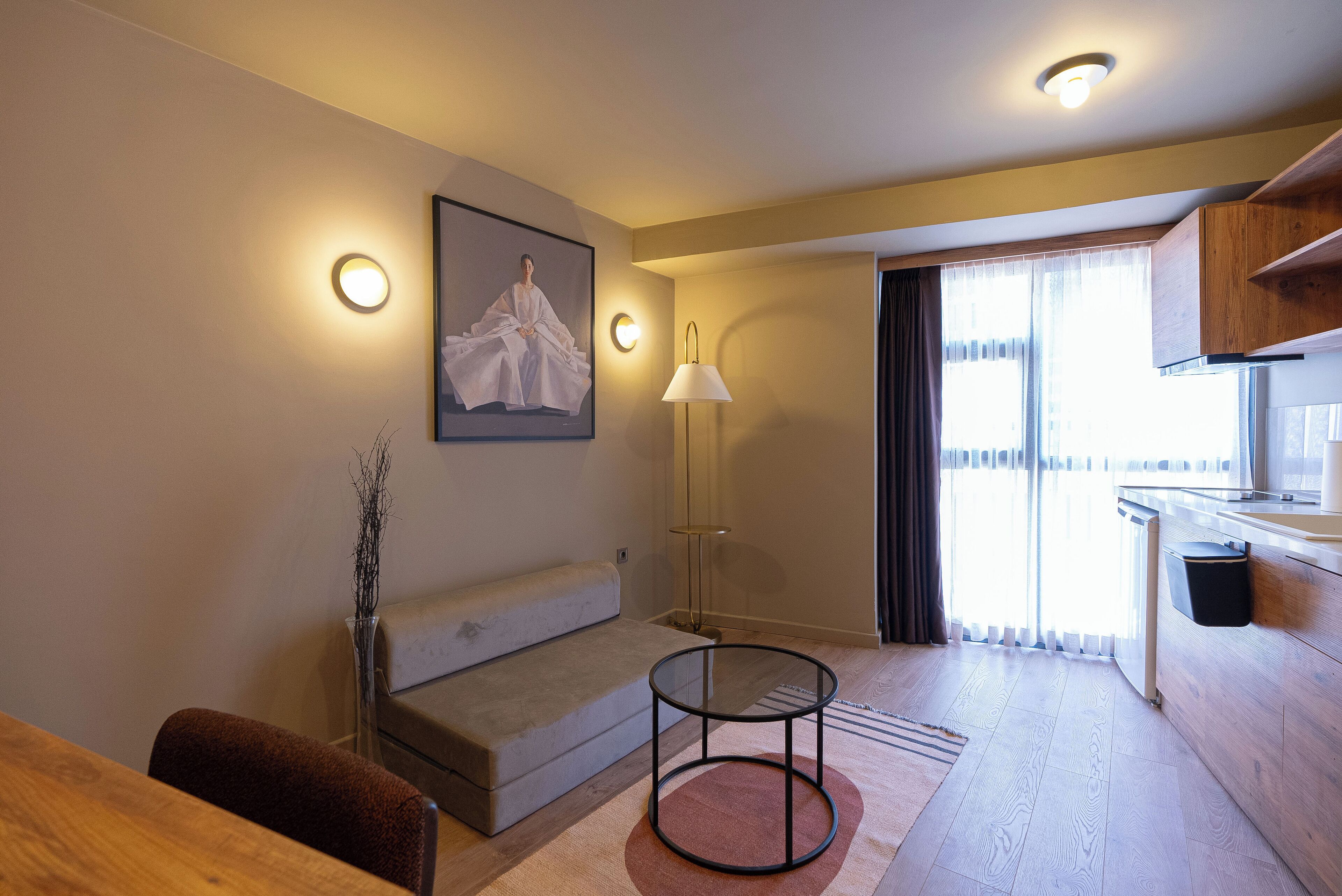 Quarto de hotel moderno com sofá, mesa redonda pequena, luminária, quadro na parede e vista para o exterior pela janela.