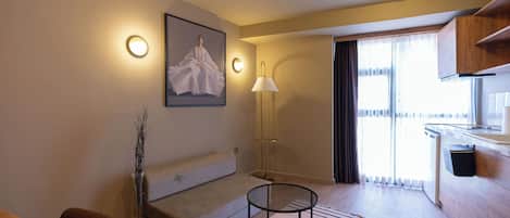 Quarto de hotel moderno com sofá, mesa redonda pequena, luminária, quadro na parede e vista para o exterior pela janela.