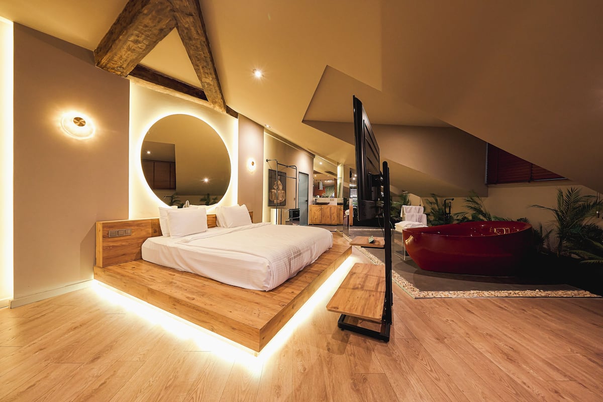 A modern bedroom with a large bed, a wooden headboard, a circular mirror, and a red bathtub.