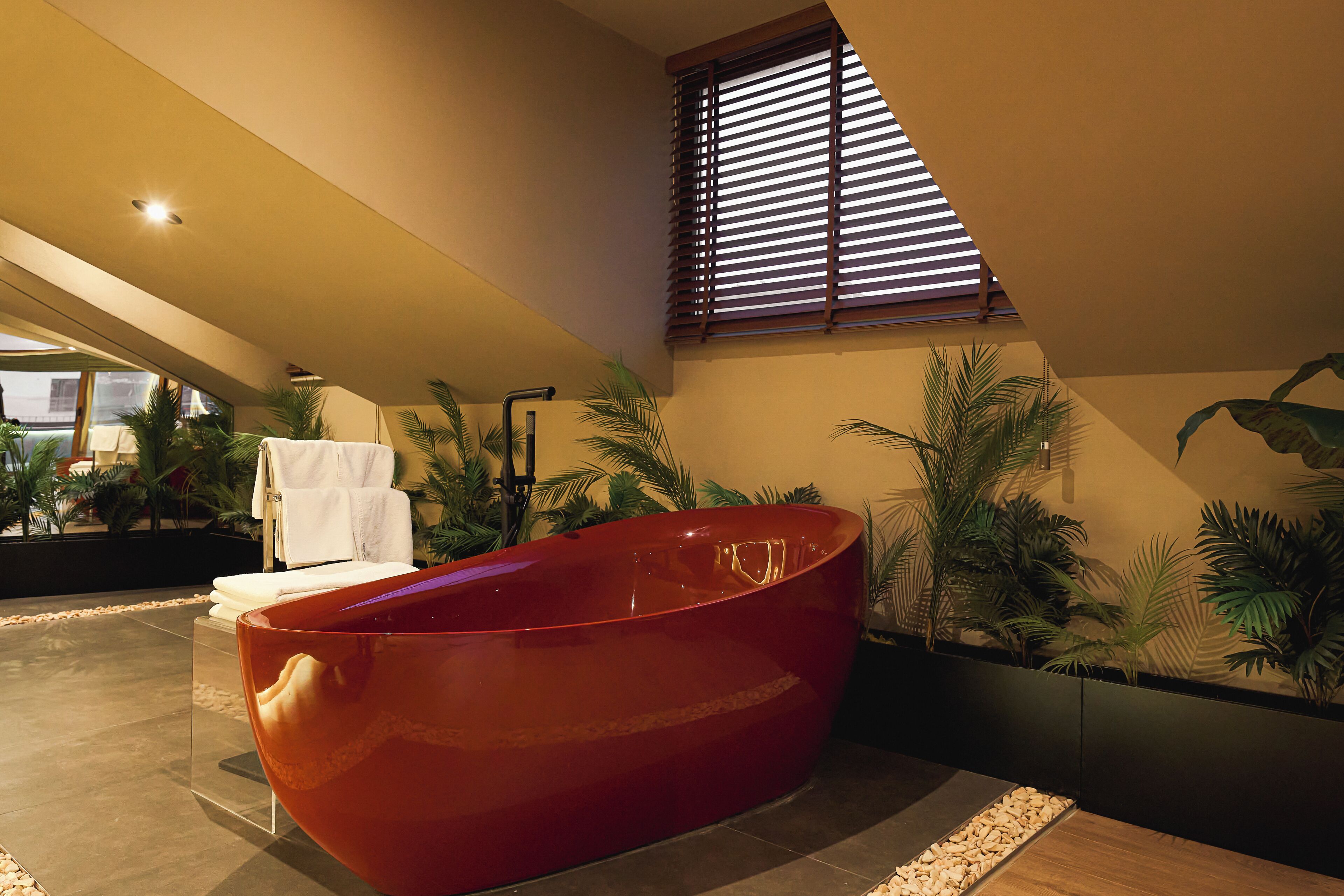 A modern bathroom with a large red bathtub, a small white sink, and a window with blinds.