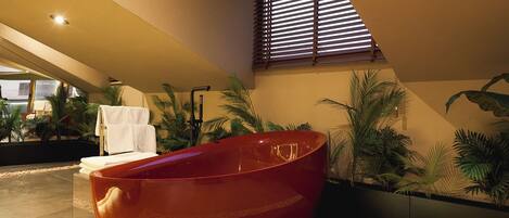A modern bathroom with a large red bathtub, a small white sink, and a window with blinds.