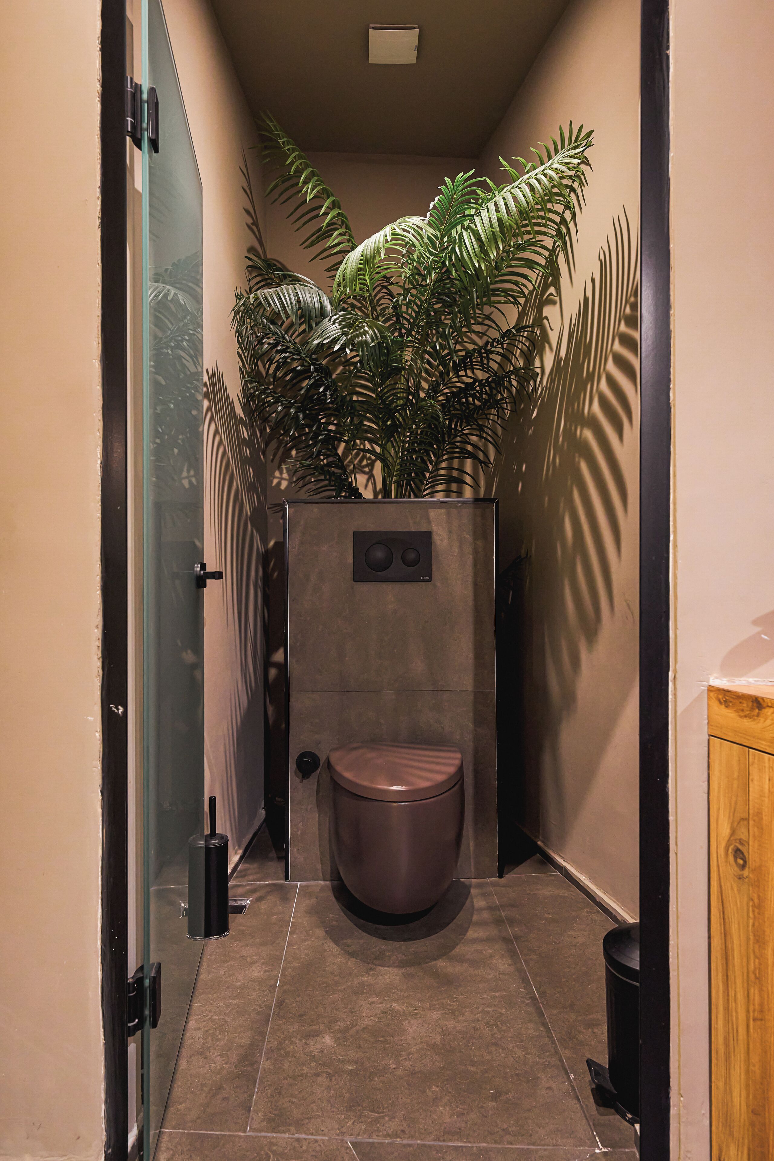 A compact bathroom with a toilet, a trash bin, and a potted plant.