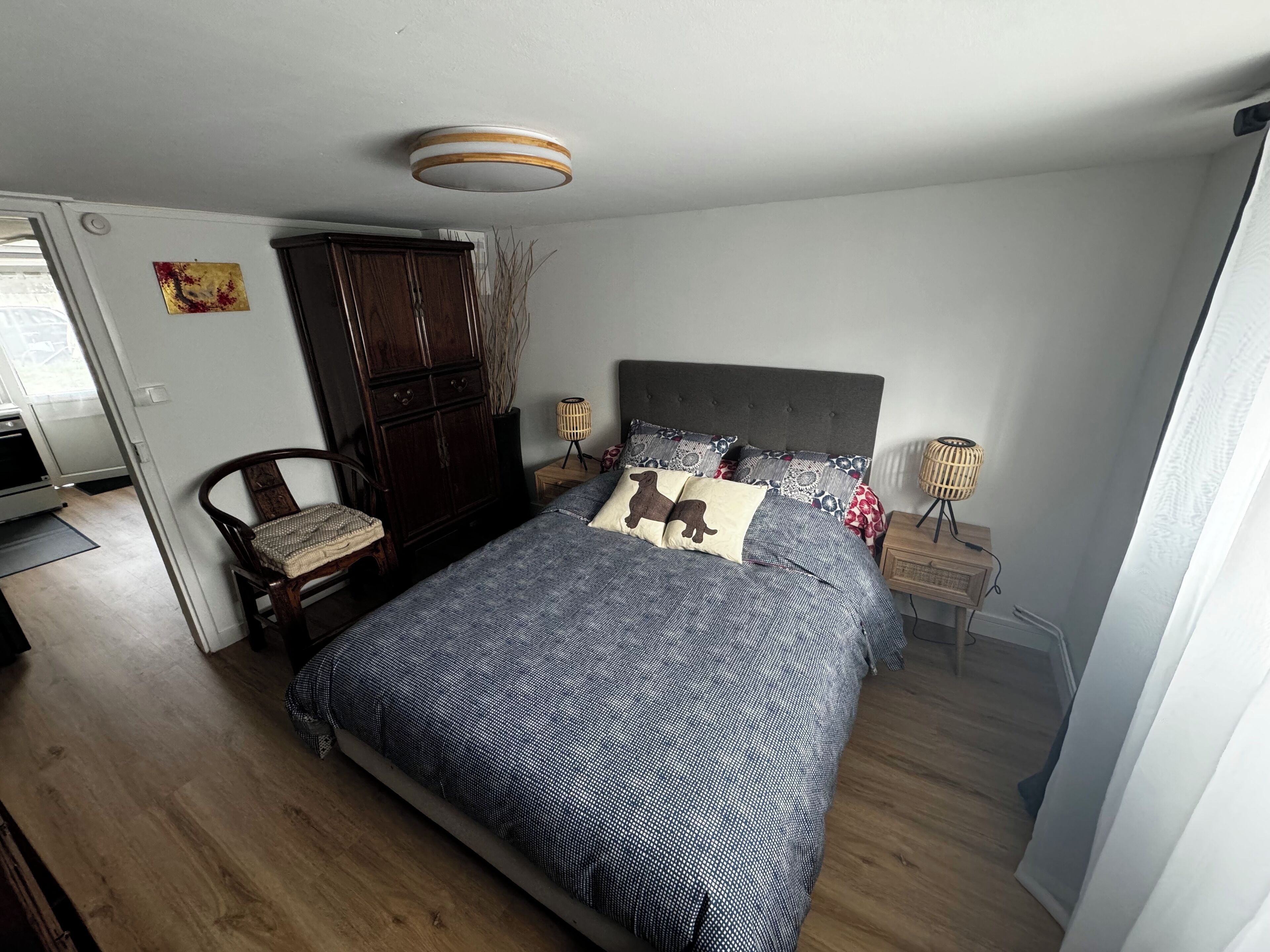 A bedroom with a large bed, a dark wooden wardrobe, a chair, a bedside table with a lamp, and a ceiling light fixture.