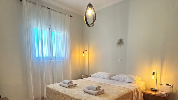 A bedroom with a bed, two bedside lamps, a nightstand, a window with curtains, and a ceiling fan. - Elli Cozy House (Corfu)