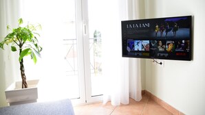 A room with a TV mounted on the wall displaying a movie, a potted plant, a sliding glass door, and a tiled floor. - Elli Cozy House (Corfu)