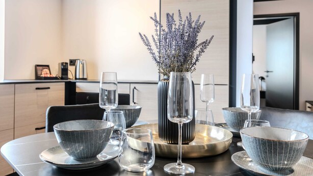 A modern dining area with a table set for two, featuring a vase with lavender, clear glasses, and grey bowls.
