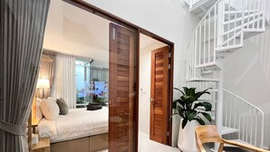 Un dormitorio con un espejo grande, una cama con almohadas, una mesita de noche y una planta en maceta. Se ve una puerta de madera que da a otra habitación y una escalera blanca.