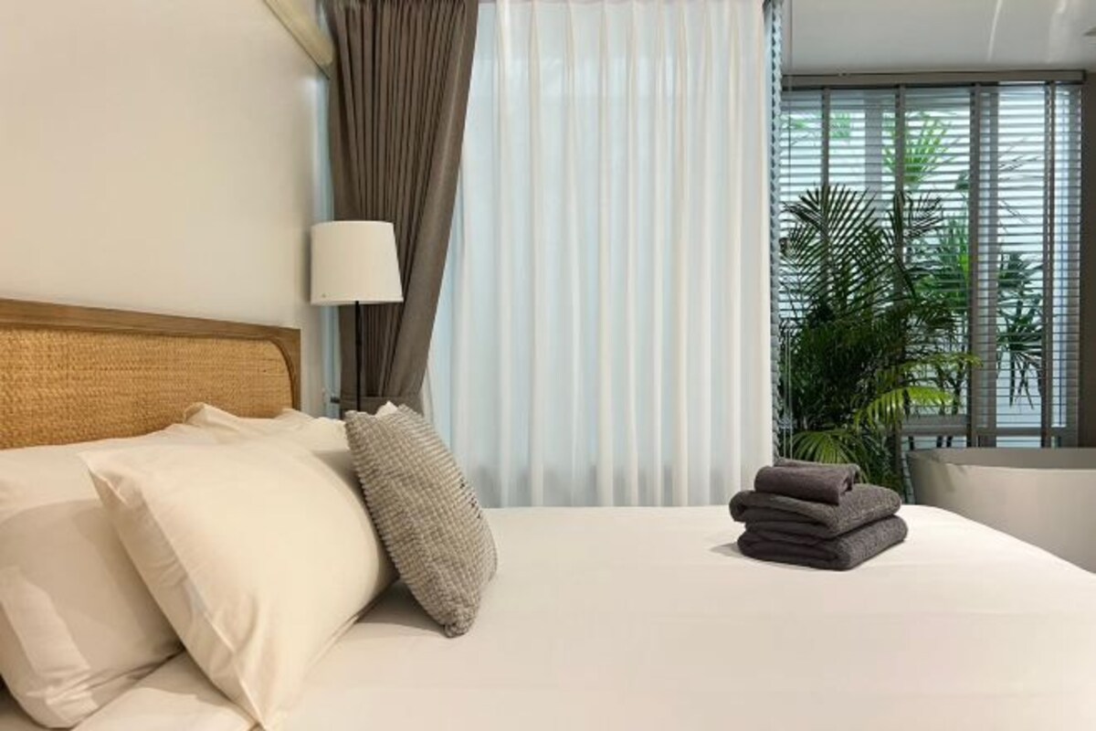 A hotel room with a bed, pillows, a lamp, curtains, and a view of palm trees through the window.