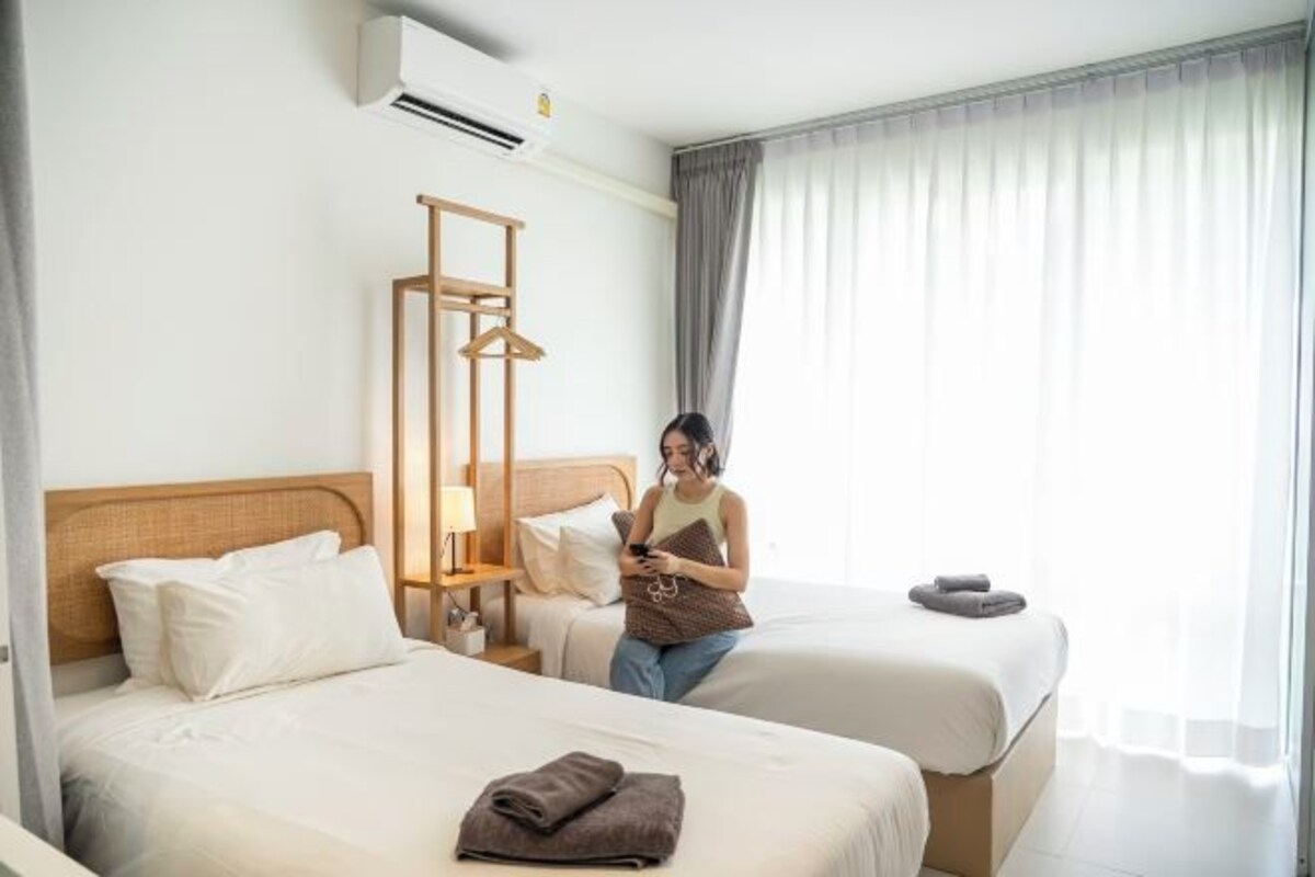 A hotel room with two beds, a wooden headboard, a hanging coat rack, and a large window with curtains.