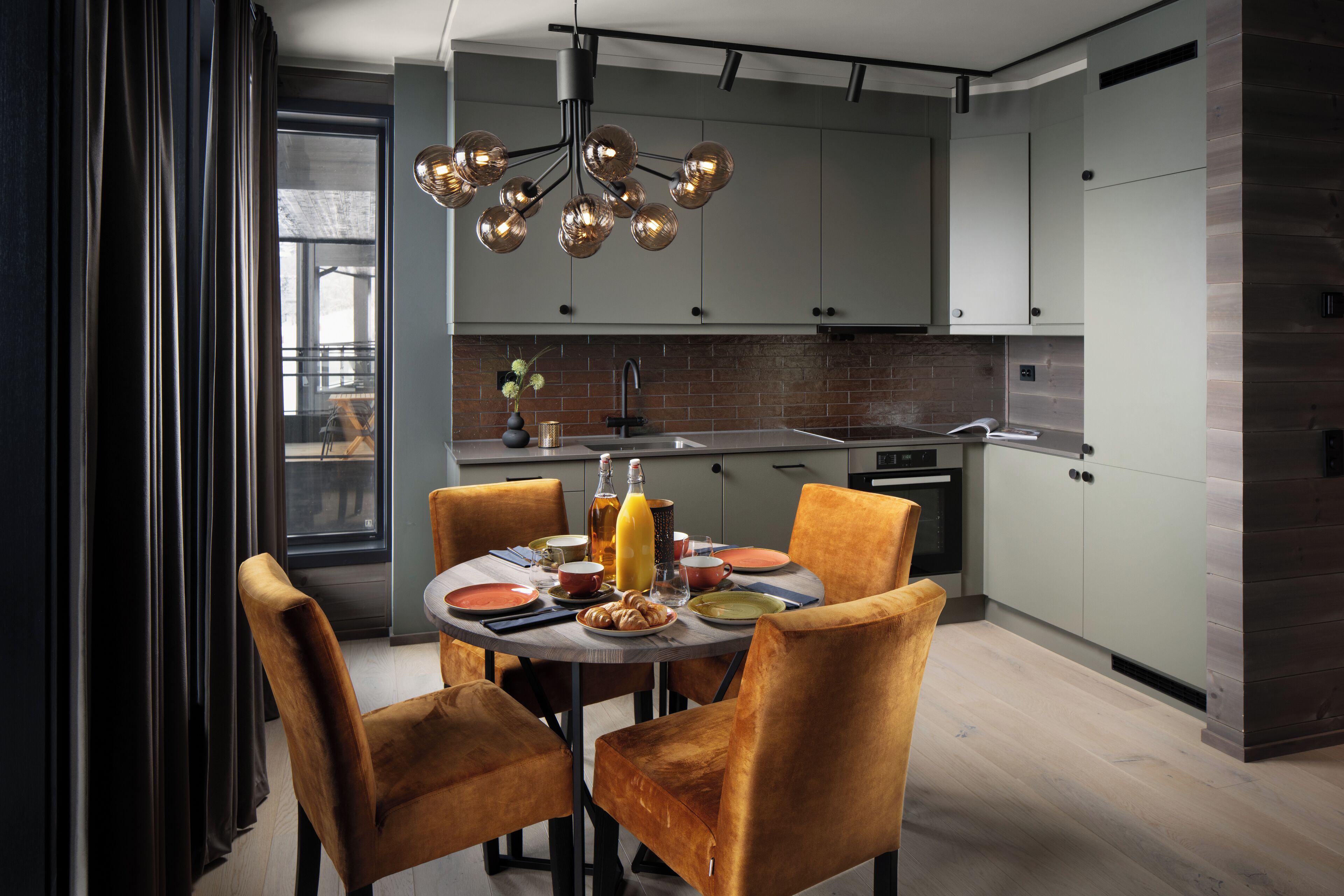 A modern kitchen with grey cabinets, a brick backsplash, and a dining area with a round table and orange chairs.