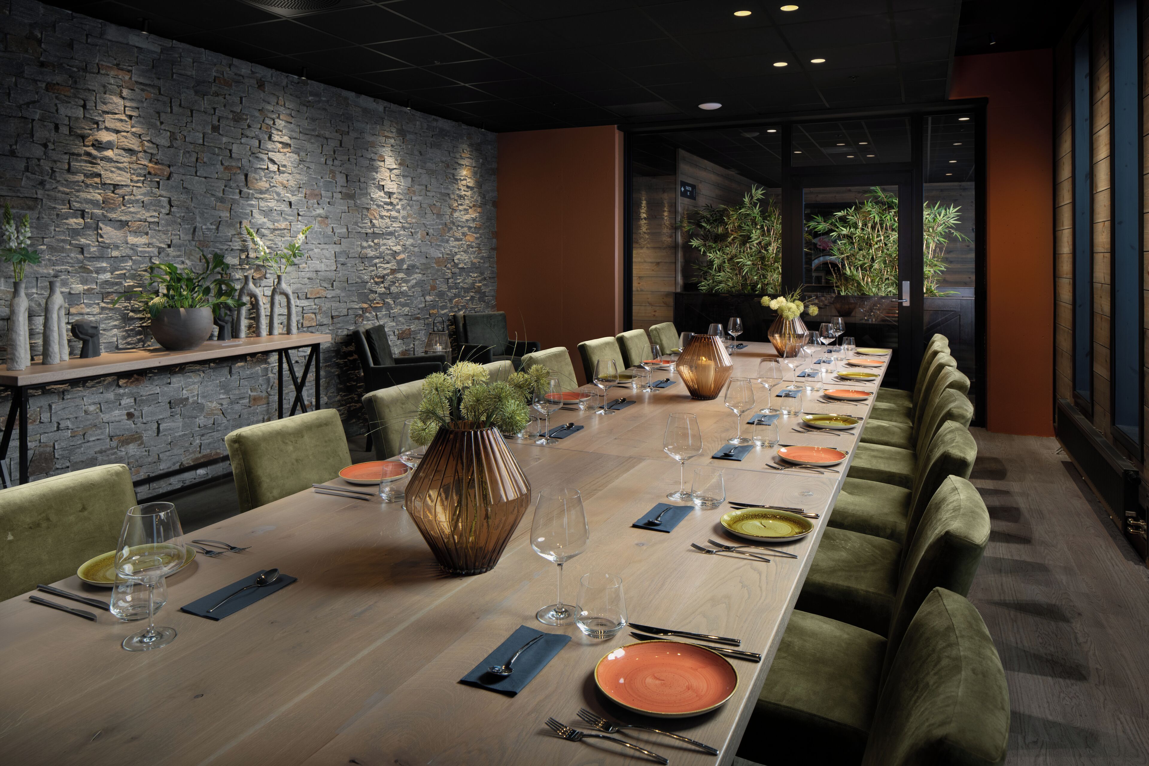 Una larga mesa de comedor dispuesta con platos, vasos y cubiertos, flanqueada por sillas tapizadas de verde, en una habitación con pared de piedra y vistas a la vegetación.
